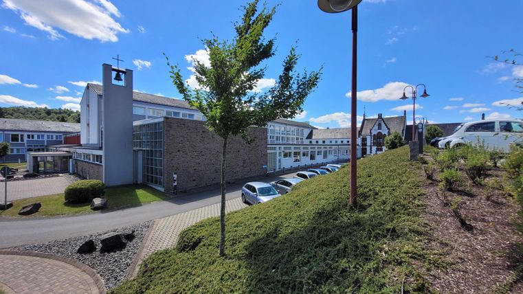 Ein modernes Gebäude mit einem großen Fensterbereich und einer Uhr auf dem Dach. Im Vordergrund gibt es eine grüne Bepflanzung und einige Autos auf dem Parkplatz.