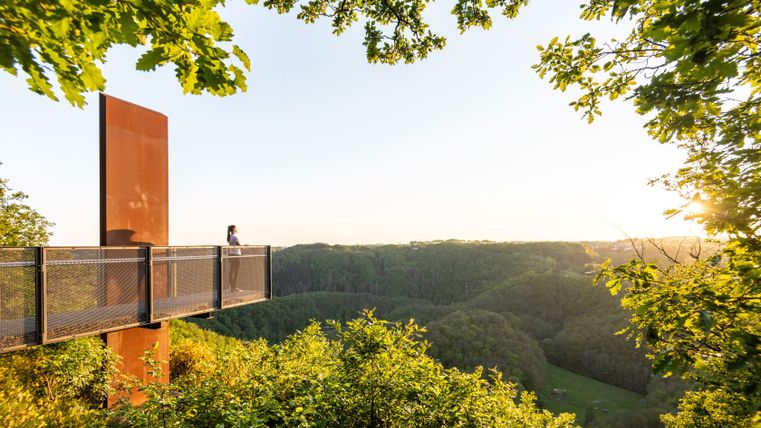 Person steht auf einem Skywalk mit Blick auf bewaldete Hügel bei Sonnenuntergang.