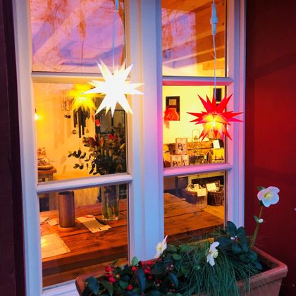 Ein gemütlicher Fensterbereich mit bunten Lichtern in Sternform. Im Inneren sind Pflanzen und eine einladende Einrichtung zu sehen.