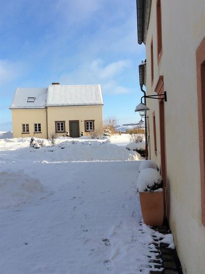 Ein schneebedeckter Innenhof mit einem kleinen gelben Haus im Hintergrund. Die Umgebung ist ruhig und winterlich mit klarem Himmel.