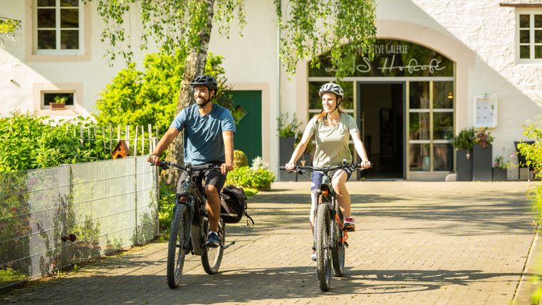 Twee mensen fietsen op een geplaveid pad voor een gebouw met de naam 'Landhofcafé'.