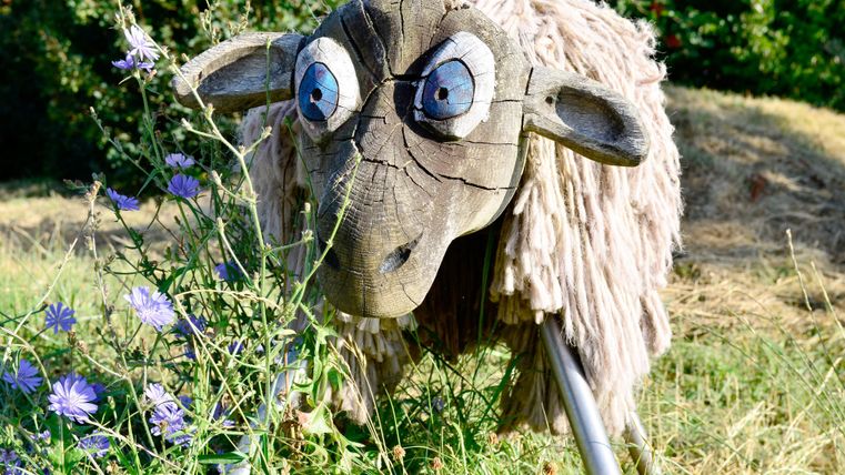 A decorative wooden sheep with a funny expression stands in the grass. It is surrounded by colorful flowers and green vegetation.