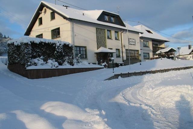 Ein Haus im Schnee mit einem gepflasterten Weg und Bäumen im Vordergrund. Der Himmel ist bewölkt und es ist ruhig und winterlich.