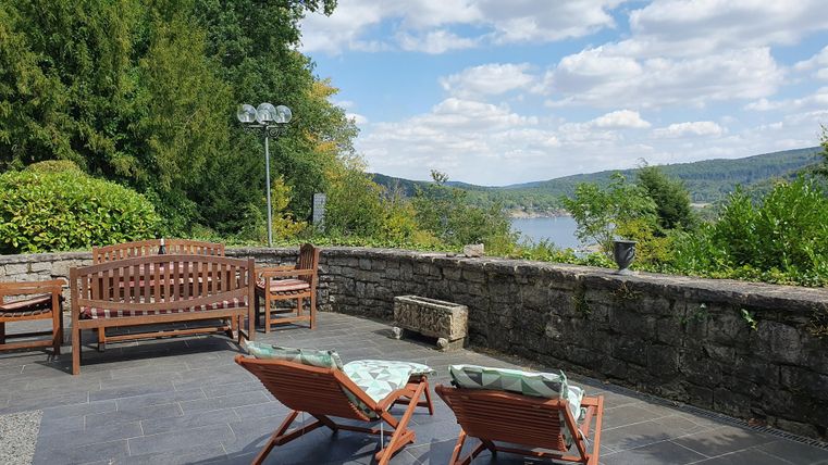 Ein gemütlicher Außenbereich mit Liegestühlen und einer schönen Aussicht auf den Fluss. Im Hintergrund sind Bäume und ein teilweise bewölkter Himmel zu sehen.