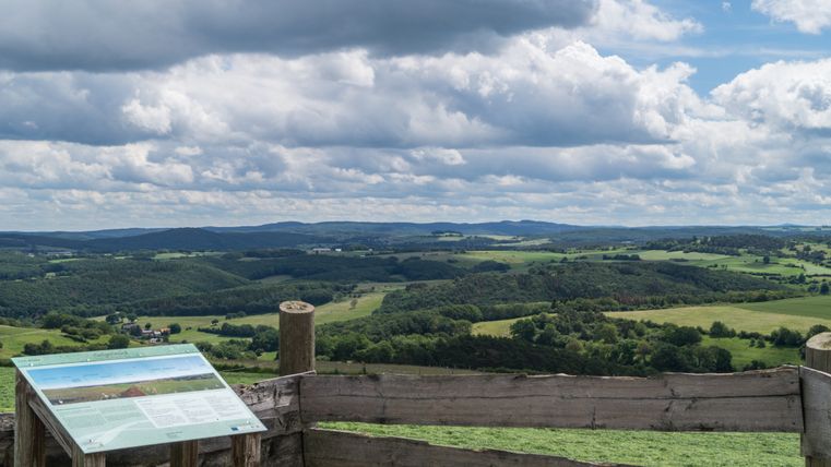 Panoramic view over the Eifel with green hills and a cloudy sky, an information sign in the foreground.