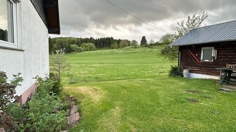 Eine grüne Wiese mit einem Blick auf bewölkten Himmel. Im Vordergrund sind zwei Gebäude, eines aus Stein und das andere aus Holz.