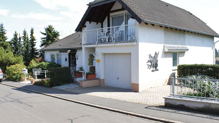Een helder eengezinswoning met een balkon en een kleine tuinlandschap. De straat is rustig, omgeven door bomen en planten.