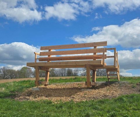 Un grand banc en bois est situé sur une pelouse sous un ciel bleu avec des nuages. Le banc offre un beau point de vue sur les environs.