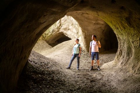 Zwei Personen stehen in einer großen, natürlichen Höhle mit erdigen Wänden und Öffnungen.