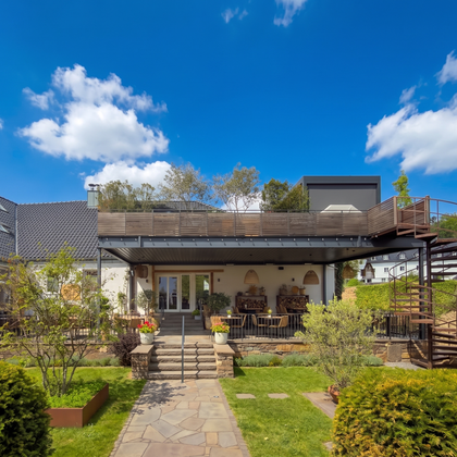A modern house with a wooden porch and a staircase leading to a rooftop garden. The garden is well-maintained with green plants and a beautiful path.