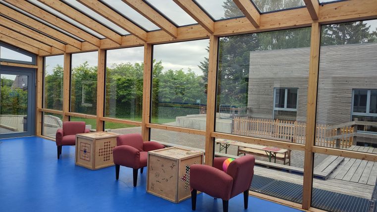 Une pièce lumineuse avec de grandes fenêtres et un sol bleu. Il y a deux chaises rouges et des tables en bois, qui créent une atmosphère accueillante.