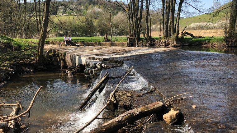 Twee mensen zitten op een bruggetje over een rivier in een landelijk landschap.