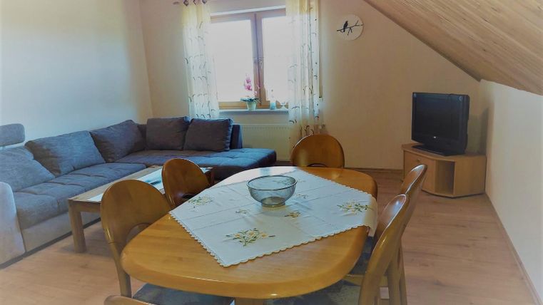 Een lichte woonkamer met een comfortabele bank en een eetgedeelte. Bij het raam staan gordijnen, en een televisie is ook aanwezig.
