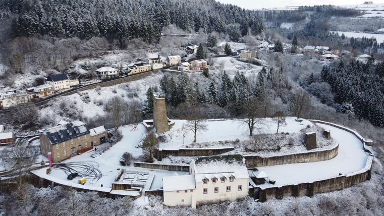 A snow-covered castle complex surrounded by a wintry landscape. In the background, the snow-covered hills and small houses are visible.
