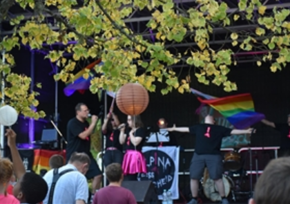 Een levendig podium met artiesten die voor een publiek zingen. Regenboogvlaggen wapperen en er heerst een feestelijke sfeer.