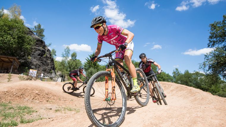 Drei Mountainbiker fahren auf einem staubigen Trail im Freien. Der Himmel ist blau mit einigen weißen Wolken.