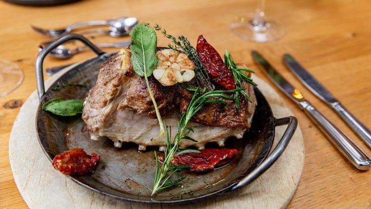 Ein saftiges Stück Fleisch auf einem servierfertigen Teller, garniert mit Kräutern und getrockneten Tomaten. Der Tisch ist aus Holz mit Besteck und weiteren Speisen im Hintergrund.