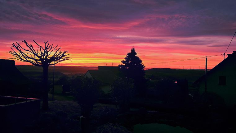 Un magnifique coucher de soleil avec des nuages roses et oranges éclatants. Des silhouettes d’arbres et de maisons confèrent à la scène une atmosphère paisible.