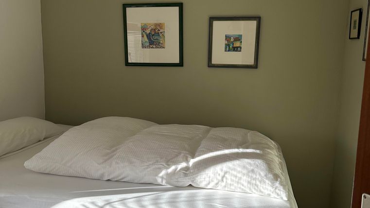 A cozy bedroom with a high bed and fresh bedding. Two pictures hang on the wall, and the light falls gently through a window.