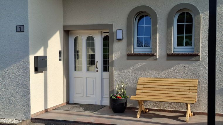 Een uitnodigende voordeur met een houten bank en een bloempot. De gevel heeft ronde ramen en een lichte kleurstelling.