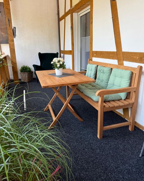 Un coin salon confortable sur un balcon avec une table en bois et un banc. Sur la table se trouve un petit vase à fleurs et en arrière-plan, des plantes sont visibles.