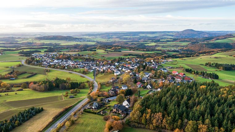 Eine malerische Landschaft mit einem kleinen Dorf und grünen Feldern. Im Hintergrund sind sanfte Hügel und Wälder zu sehen.
