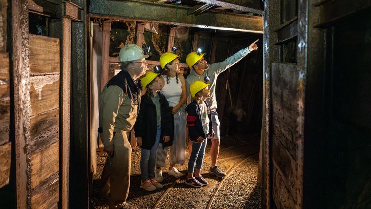 Eine Gruppe von fünf Personen in einem Bergwerk schaut neugierig nach vorne. Sie tragen Schutzhelme und stehen in einem beleuchteten Tunnel.
