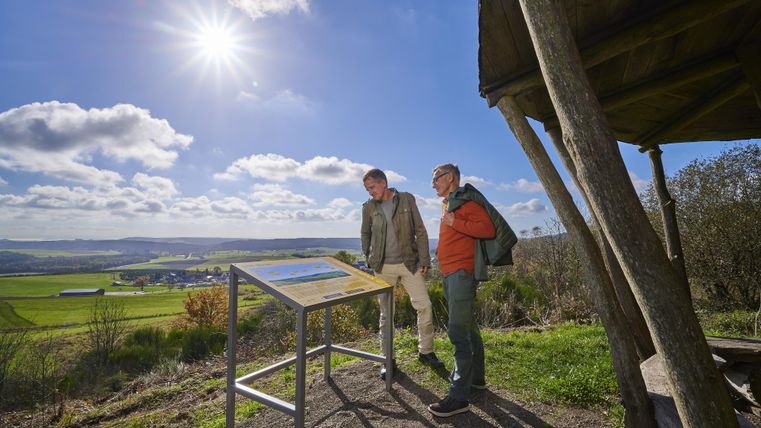 Two people are standing at an information board in nature. In the background, a green landscape is visible under a clear sky.