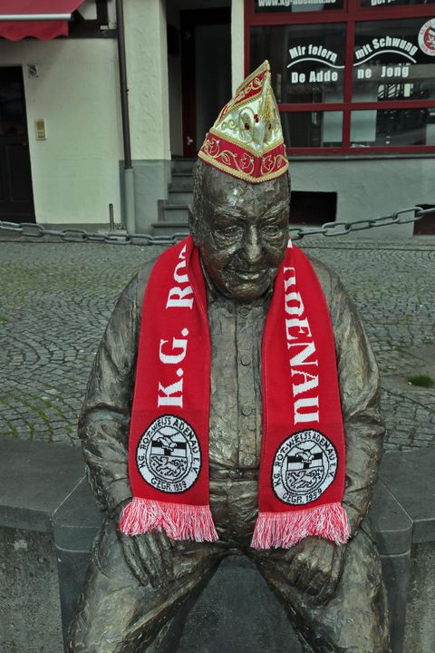 A bronze statue of a seated man wearing a red scarf and a festive hat. The statue is placed in a cobblestone square in the city.