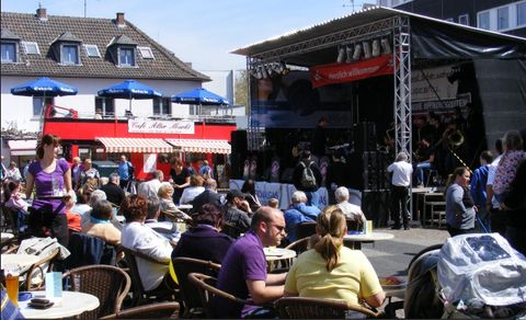 Ein lebhaftes Festival mit einer Bühne und einer Band, die spielt. Viele Menschen sitzen an Tischen und genießen die Musik im Freien.