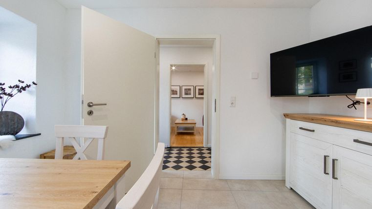 Ein modernes Wohnzimmer mit Holztisch und weißen Möbeln. Der Raum hat eine helle Atmosphäre und führt zu einem weiteren Raum mit einem freundlichen Bodenmuster.