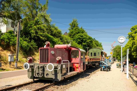 An old red locomotive is standing at a station with a carriage. In the background, there are trees and a clock visible.
