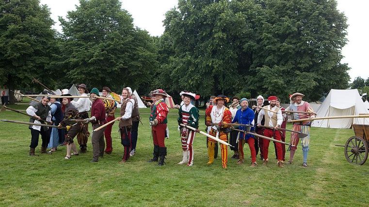 Een groep mensen in historische kostuums staat buiten. Ze houden lange stokken vast en staan voor tenten in een groen veld.