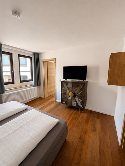 Een moderne kamer met twee bedden en een televisie aan de muur. De houten vloer en de ramen zorgen voor een heldere en uitnodigende sfeer.