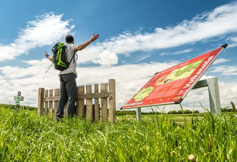 Un randonneur se tient debout, les bras écartés, devant une clôture en bois sur le sentier Vulcano. Un panneau d'information est visible au premier plan.