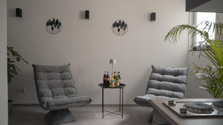 Een gezellige kamer met twee grijze fauteuils en een tafeltje. Aan de muur hangen decoratieve schilderijen en in de hoek staat een plant.