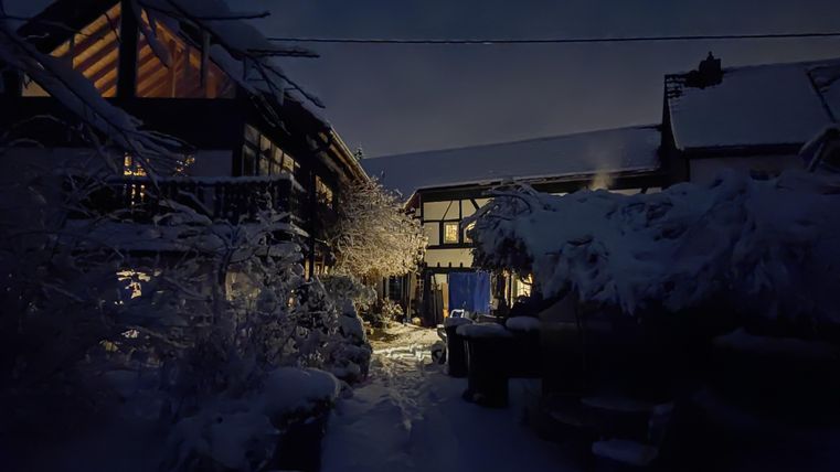 Une ferme d'hiver la nuit, avec des bâtiments recouverts de neige et un éclairage doux. Le chemin est dégagé et mène à une entrée chaleureusement illuminée.