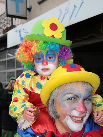 A cheerful clown carries a child on his shoulders. Both are wearing colorful costumes and have painted faces.