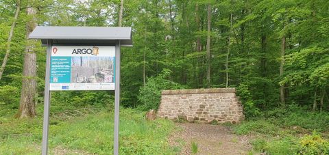 Eine Informationstafel vor einer Steinmauer inmitten eines Waldes