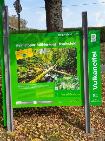 Un panneau d'information sur le HeimatSpur Mühlenweg Deudesfeld. En arrière-plan, on peut voir des arbres et un environnement paisible.