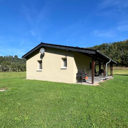 Une maison simple sur un terrain vert. À l'arrière-plan, on peut voir des arbres et un ciel bleu.