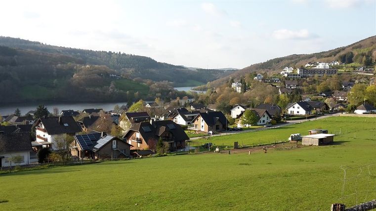Een pittoresk landschap met kleine huizen en zachte heuvels. Op de achtergrond is een rustige rivier te zien, omringd door bomen.