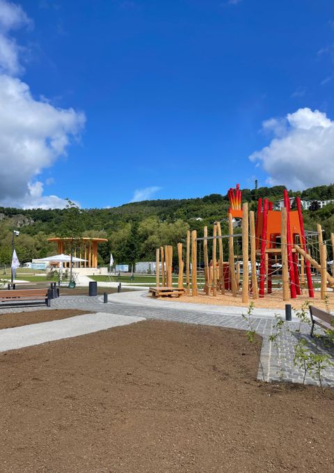 Un nouveau terrain de jeu en bois avec des éléments rouges est situé sur une surface pavée. À l'arrière-plan, on peut voir des arbres et une colline boisée.