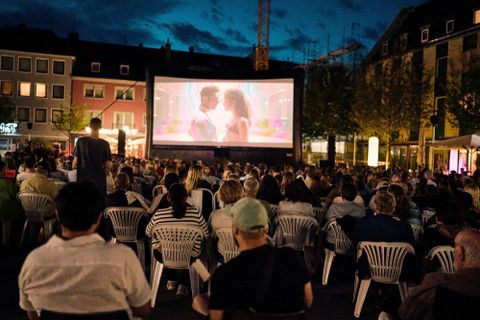 Ein Freiluftkino mit einer großen Leinwand, auf der ein Film gezeigt wird. Viele Menschen sitzen auf weißen Stühlen und genießen die Vorstellung bei Nacht.
