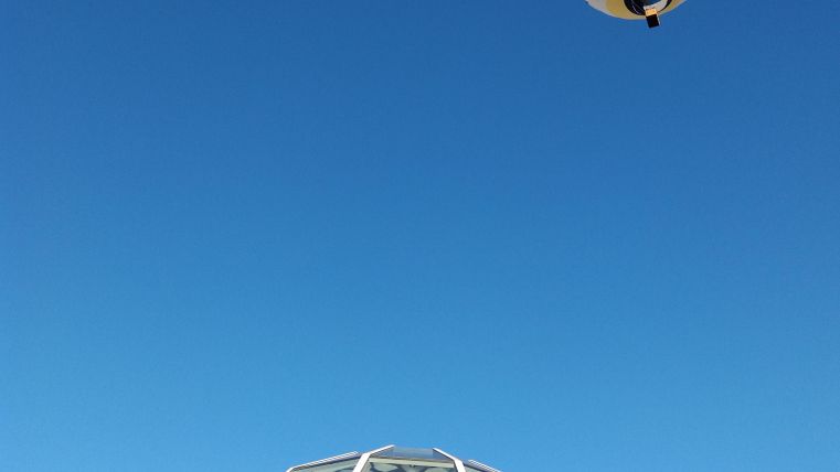 Een heteluchtballon zweeft hoog aan de hemel boven een modern gebouw. De heldere blauwe lucht vormt een mooie achtergrond.
