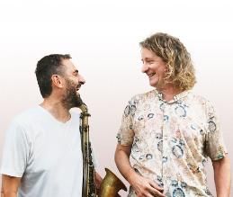Two men are standing side by side, smiling. One is holding a saxophone and they appear friendly and relaxed.