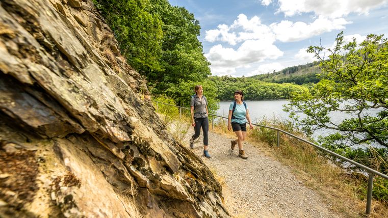Zwei Frauen wandern auf einem Pfad entlang eines Sees, umgeben von Bäumen und Felsen.