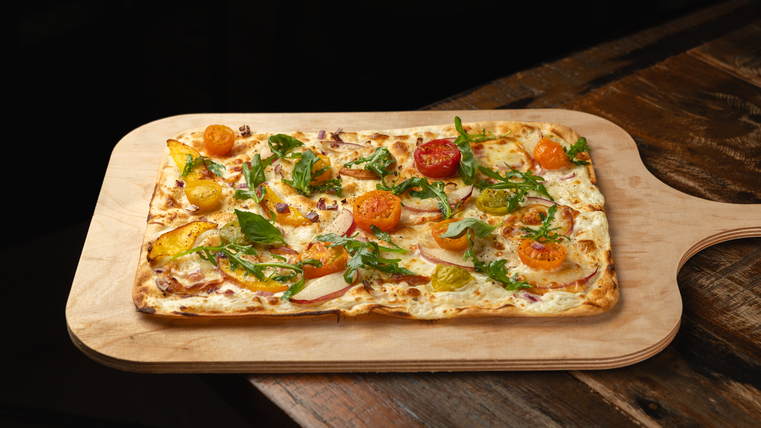 Une tarte flambée fraîchement cuite sur une planche en bois. Elle est garnie de tomates bio régionales colorées de Theo Frings et de roquette.