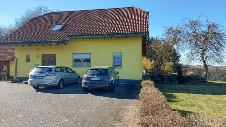 Een geel huis met een rood dak staat in een zonnige tuin. Twee auto's parkeren voor het huis, omringd door groene gazons.