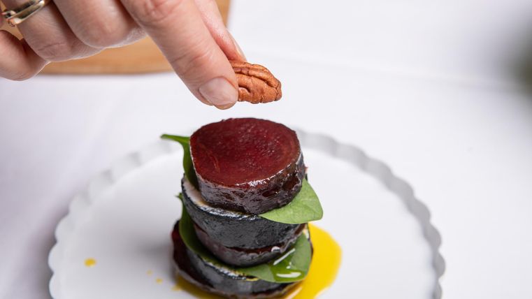 Een kunstzinnig gepresenteerd bord met lagen van gerechten, gegarneerd met een blad en een noot. Een hand voegt net een noot toe.